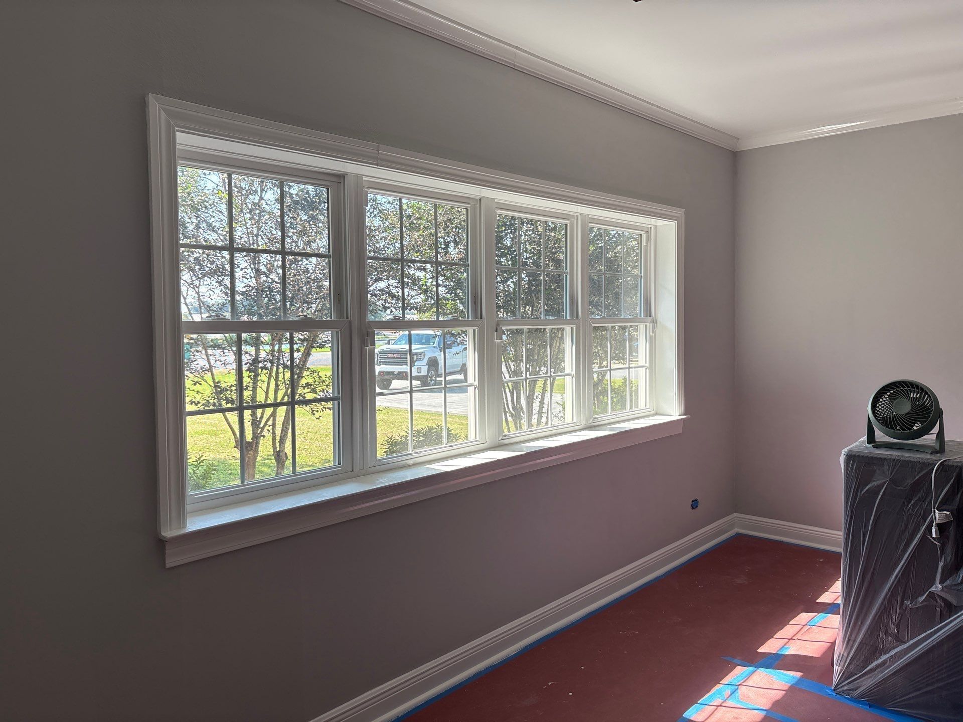 A window with white trim, gray walls, and red floor; a small fan sits on a patterned pedestal.