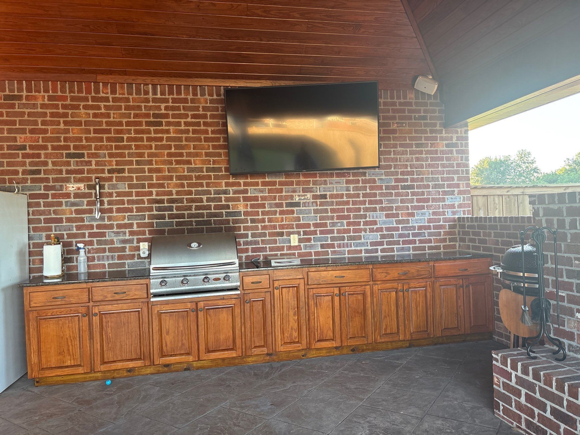 Outdoor kitchen with brick walls, wooden cabinets, a grill, and a mounted TV.