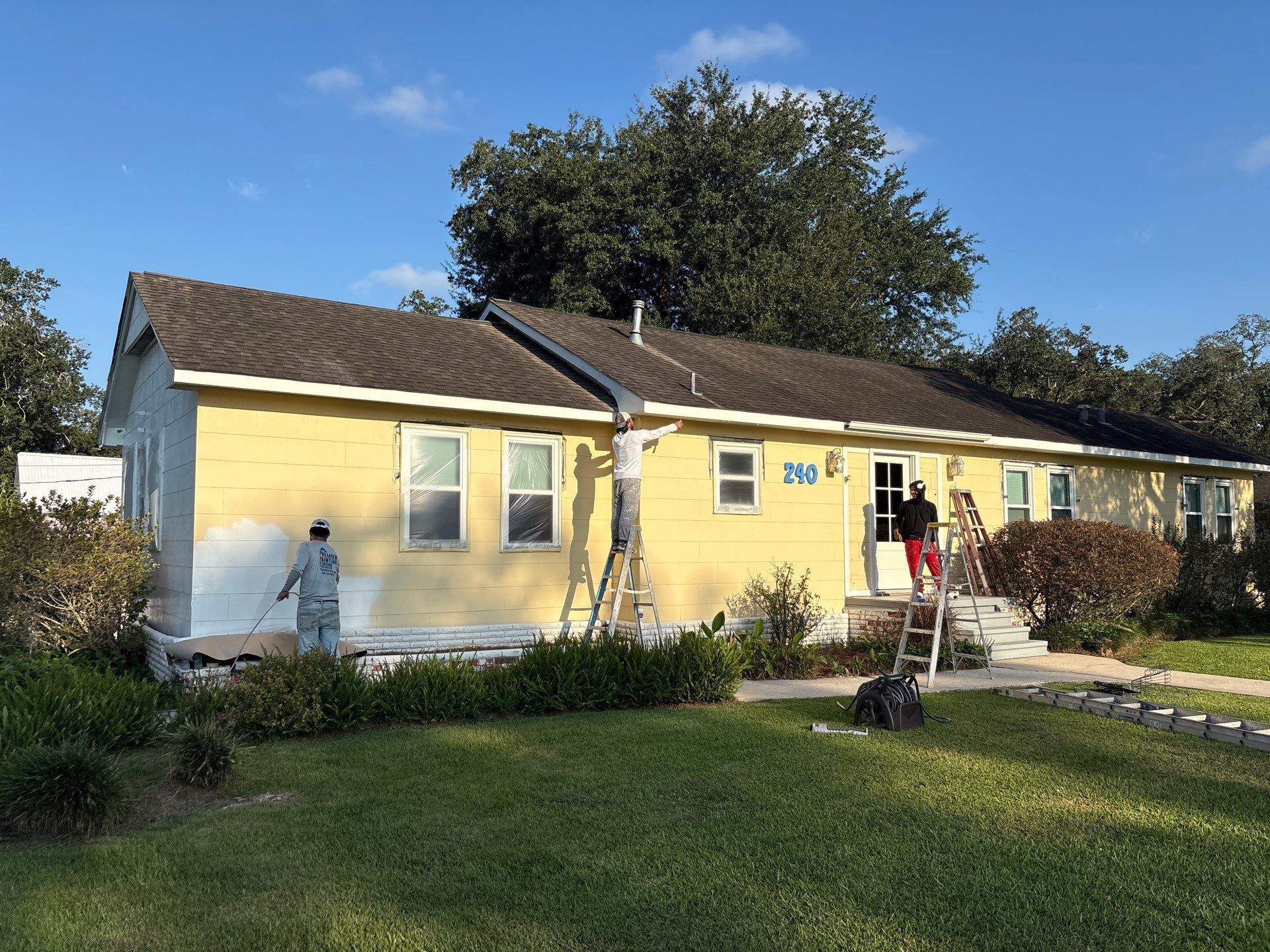 House being painted yellow; people on ladders painting the exterior. Green lawn, blue sky.