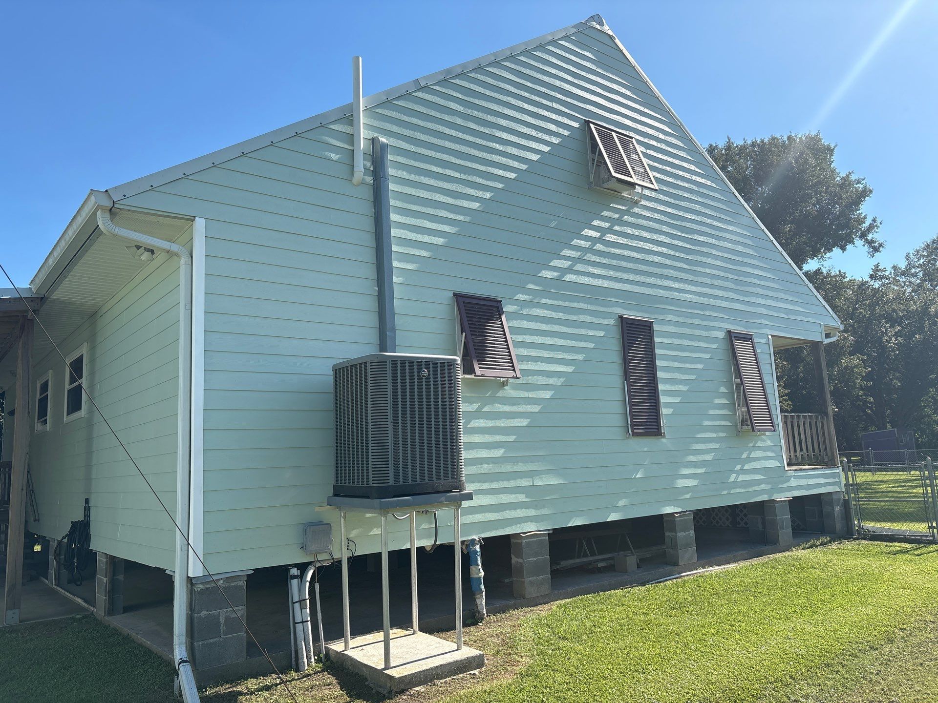 Green house with brown shutters, raised on pillars, against a blue sky, air conditioner on the side.