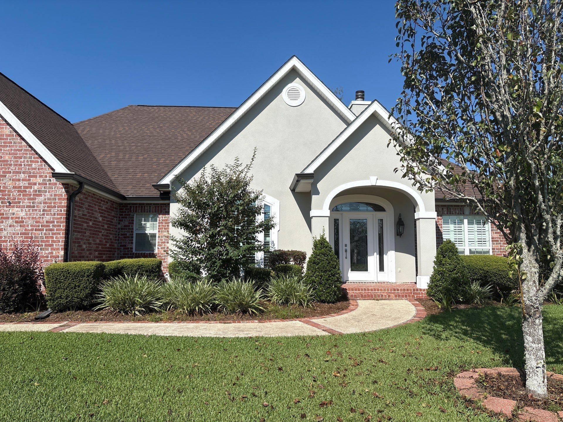 Tan stucco and brick house with arched entrance, brown roof, and green lawn.