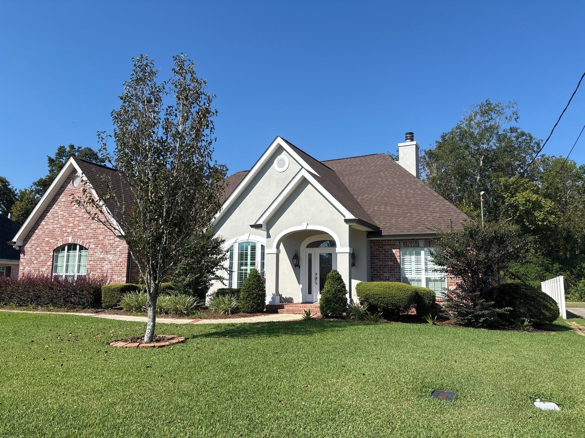 A single-story house with a brick facade on the left and stucco on the right, surrounded by green grass and bushes, under a blue sky.