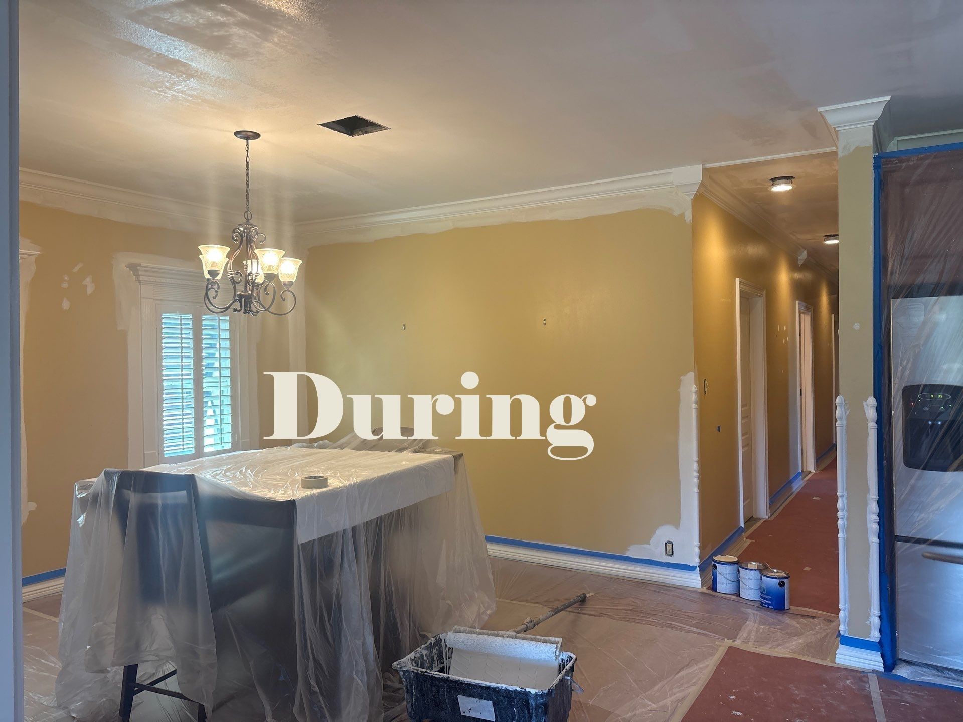 Interior during painting: kitchen with covered island, tan walls, white trim, hallway in background.