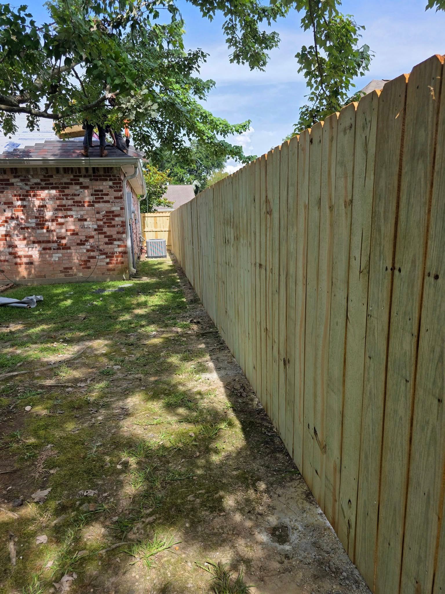 Narrow grassy yard beside a weathered wooden fence and brick building under trees