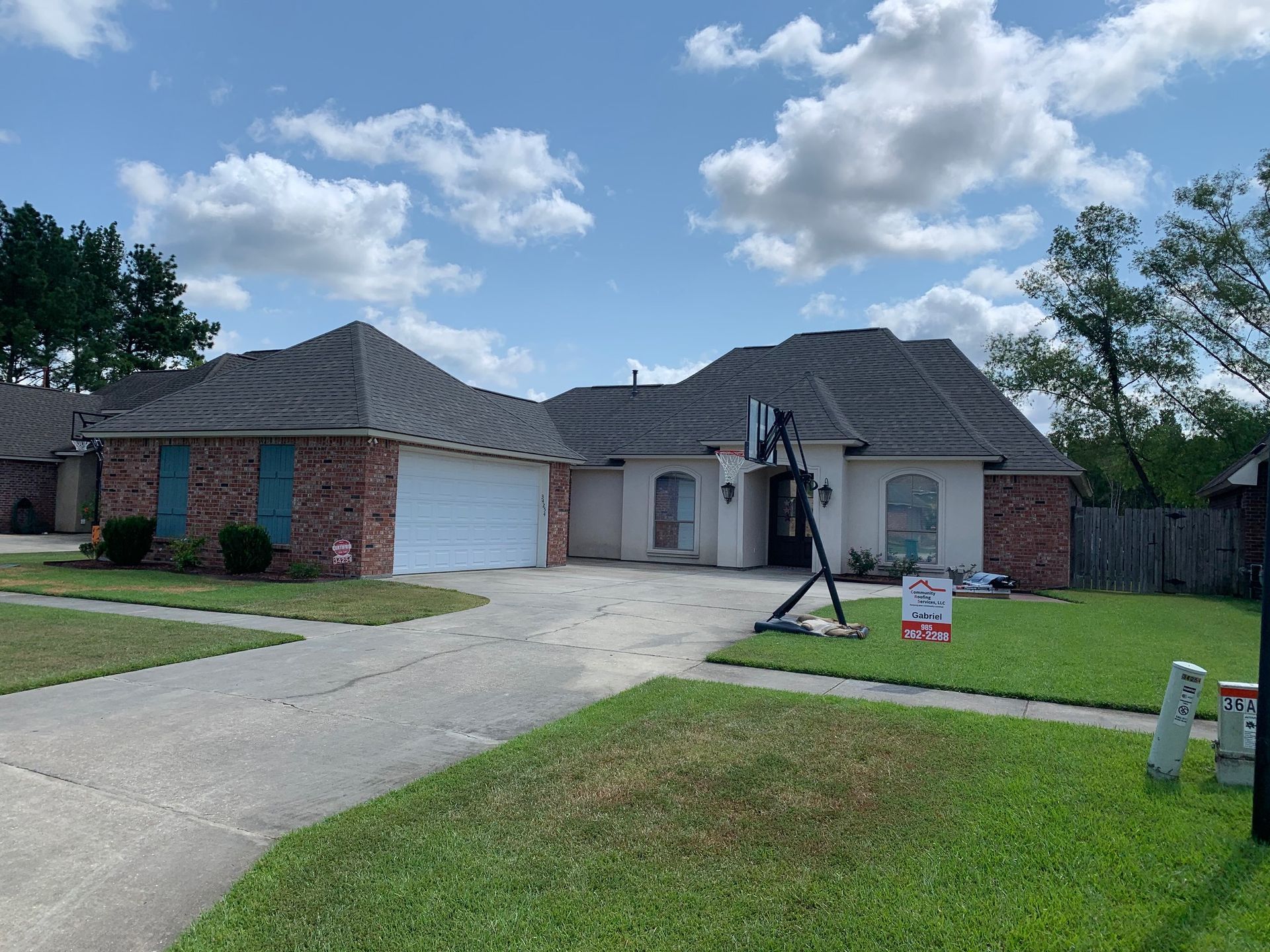 Suburban brick house with two-car garage, driveway, and a person holding a camera tripod in the front yard