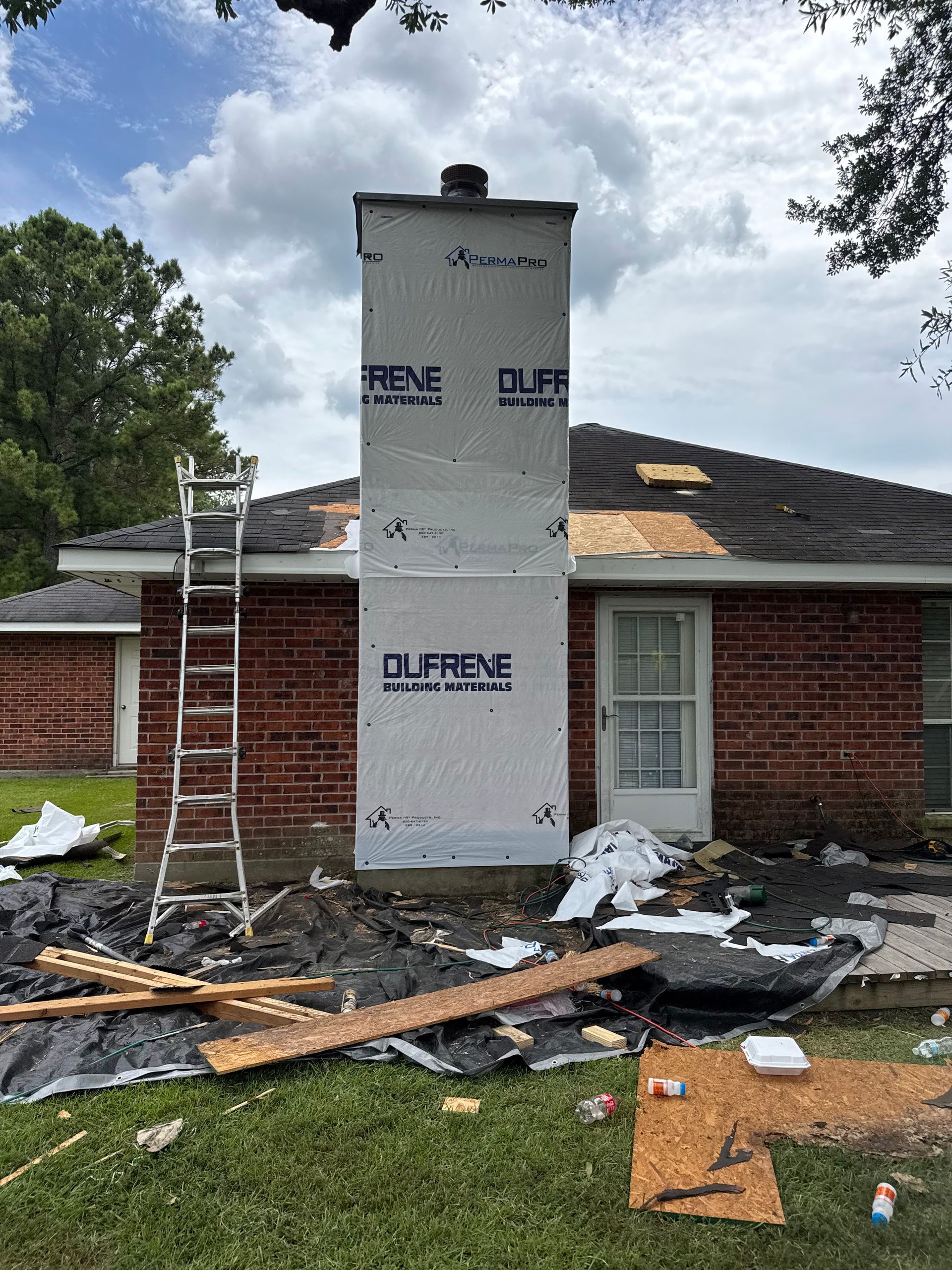 Roof under repair with new chimney wrapped in white insulation, debris and ladders in front of a brick house
