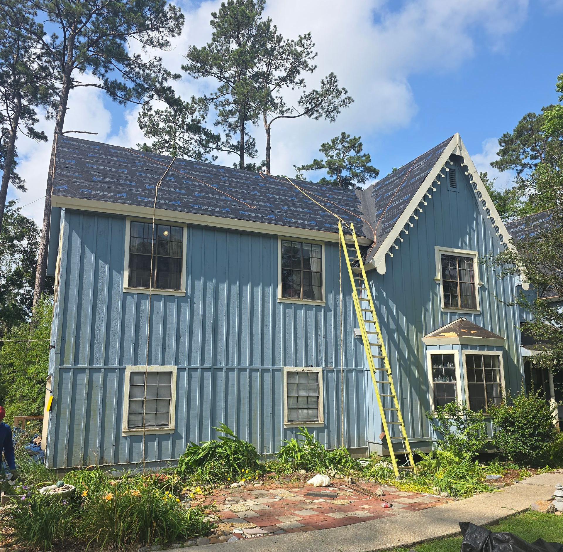 Blue two-story house with white trim and a ladder leaning against it, surrounded by trees and greenery.