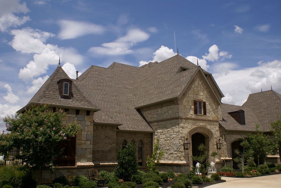 Large stone house with a steep shingle roof, arched entryway, and landscaped front yard under a partly cloudy sky
