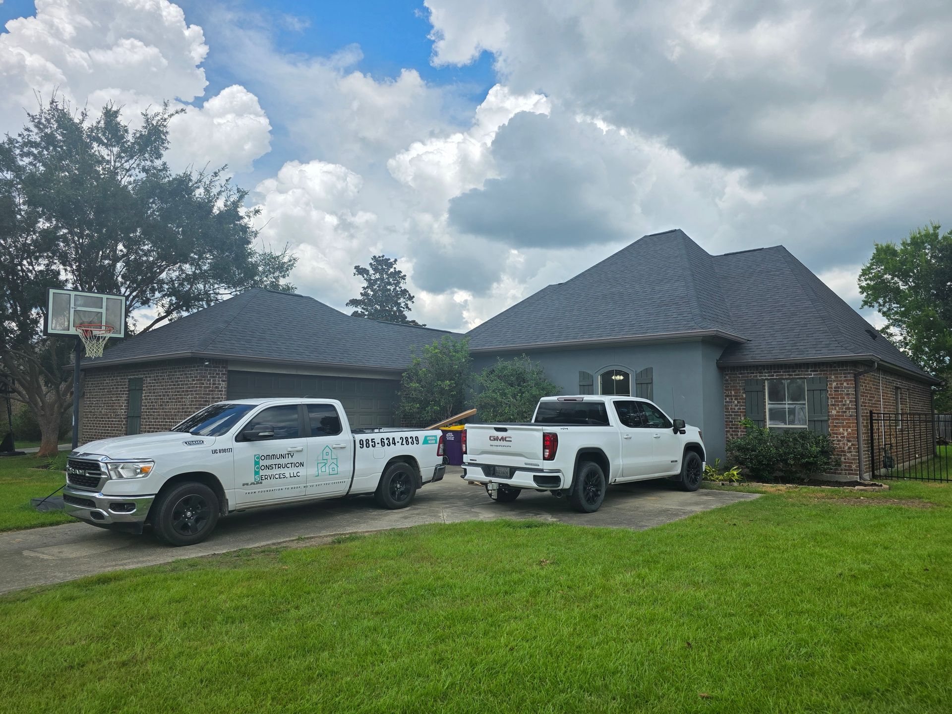 Two white pickup trucks parked outside a brick house with a dark roof on a cloudy day