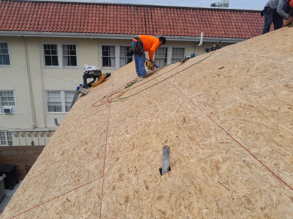 Workers installing plywood sheathing on a steep roof at a building site