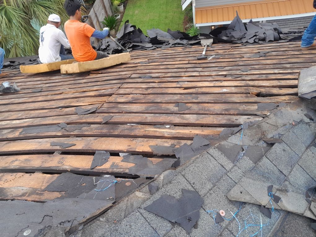 Roofers replacing dark shingles on a damaged roof, with workers kneeling near the ridge.