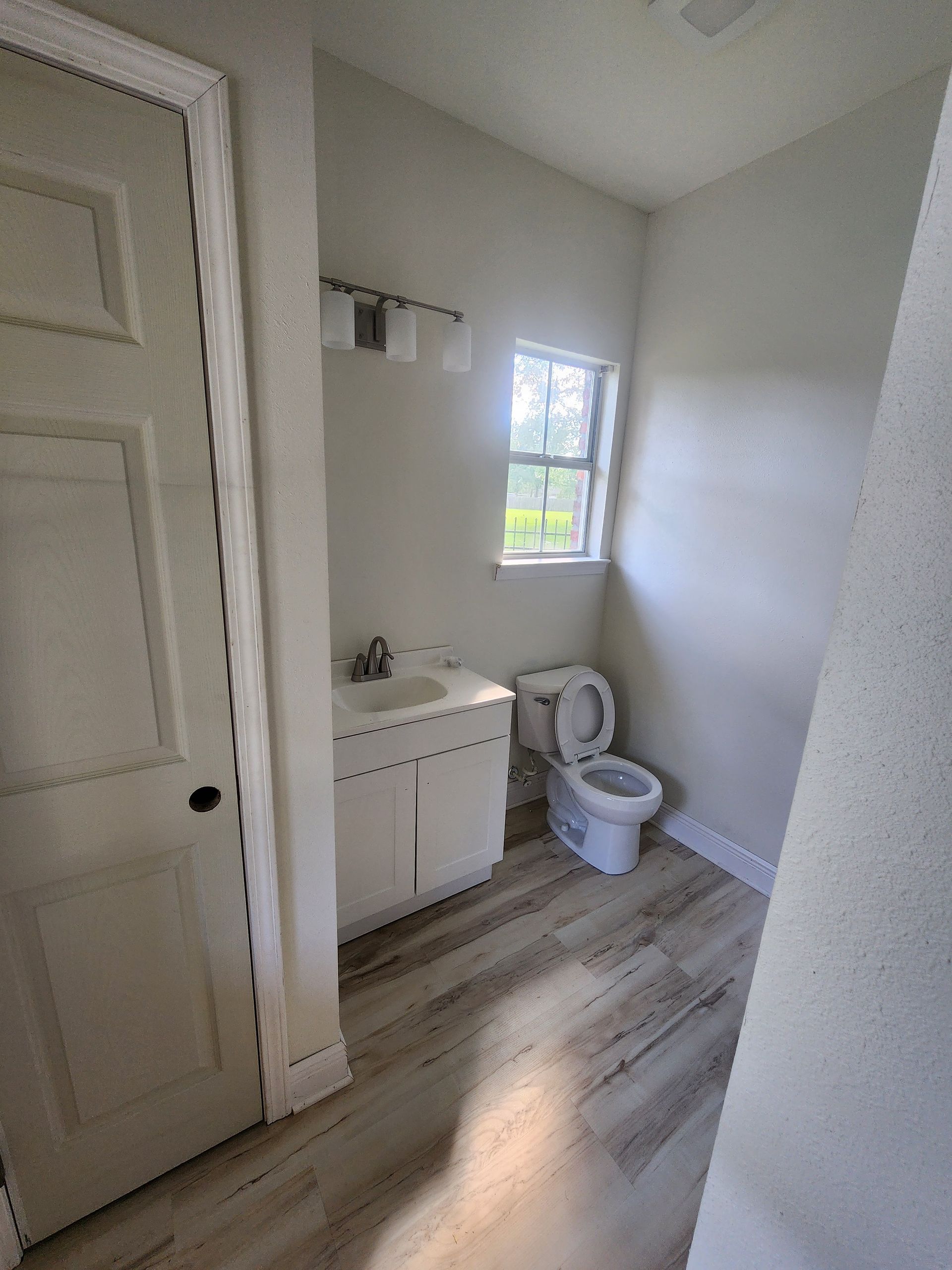 Small bathroom with white sink, toilet, window, and light wood flooring.