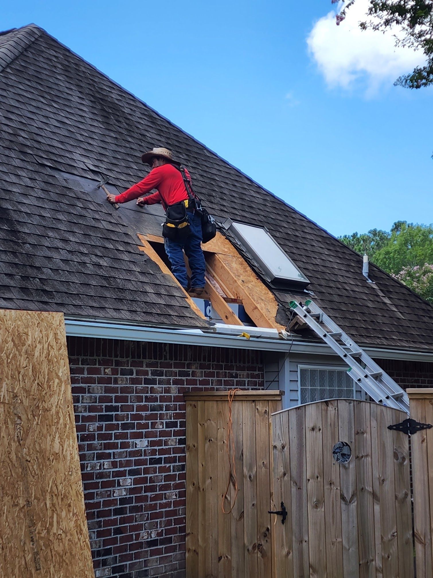 Worker on a roof installing shingles beside a ladder on a brick house