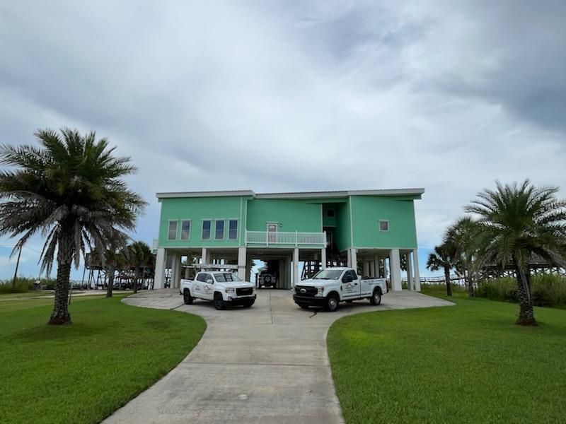 Mint-green two-story house with palm trees, parked trucks, and a cloudy sky over a curved driveway