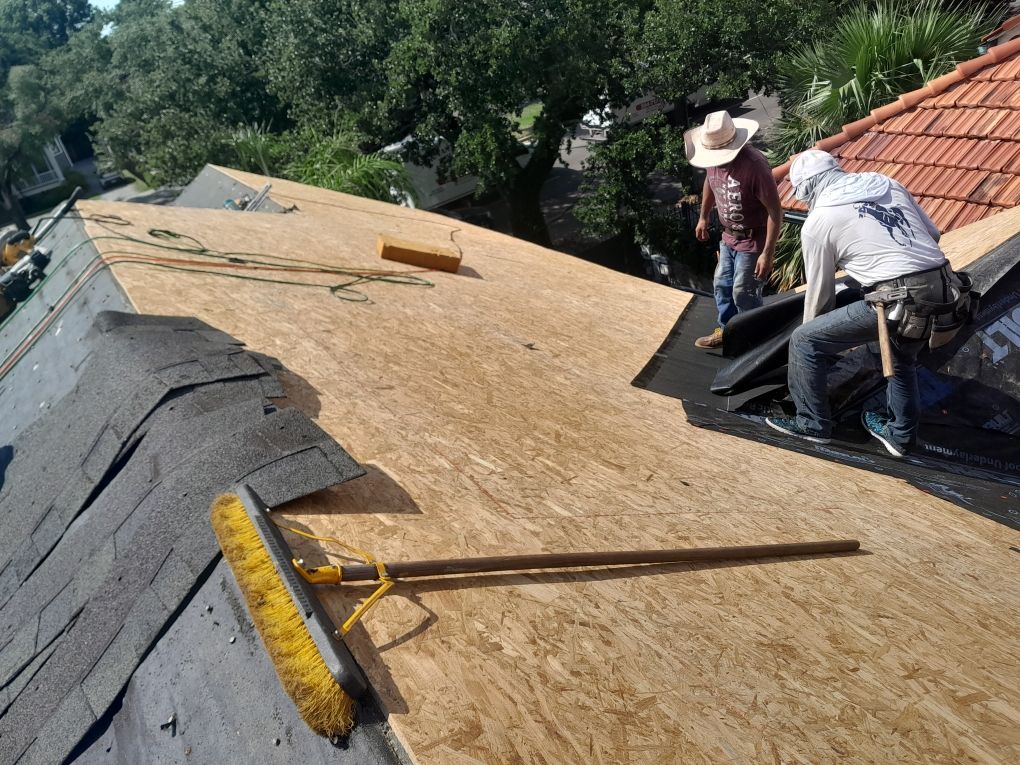 Workers installing plywood roof sheathing on a sloped roof with tools and shingles nearby