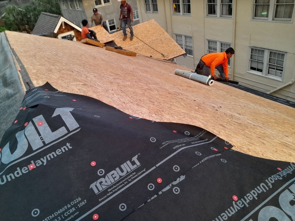 Workers installing roofing underlayment on a house roof, with orange boards and black waterproof membrane visible.