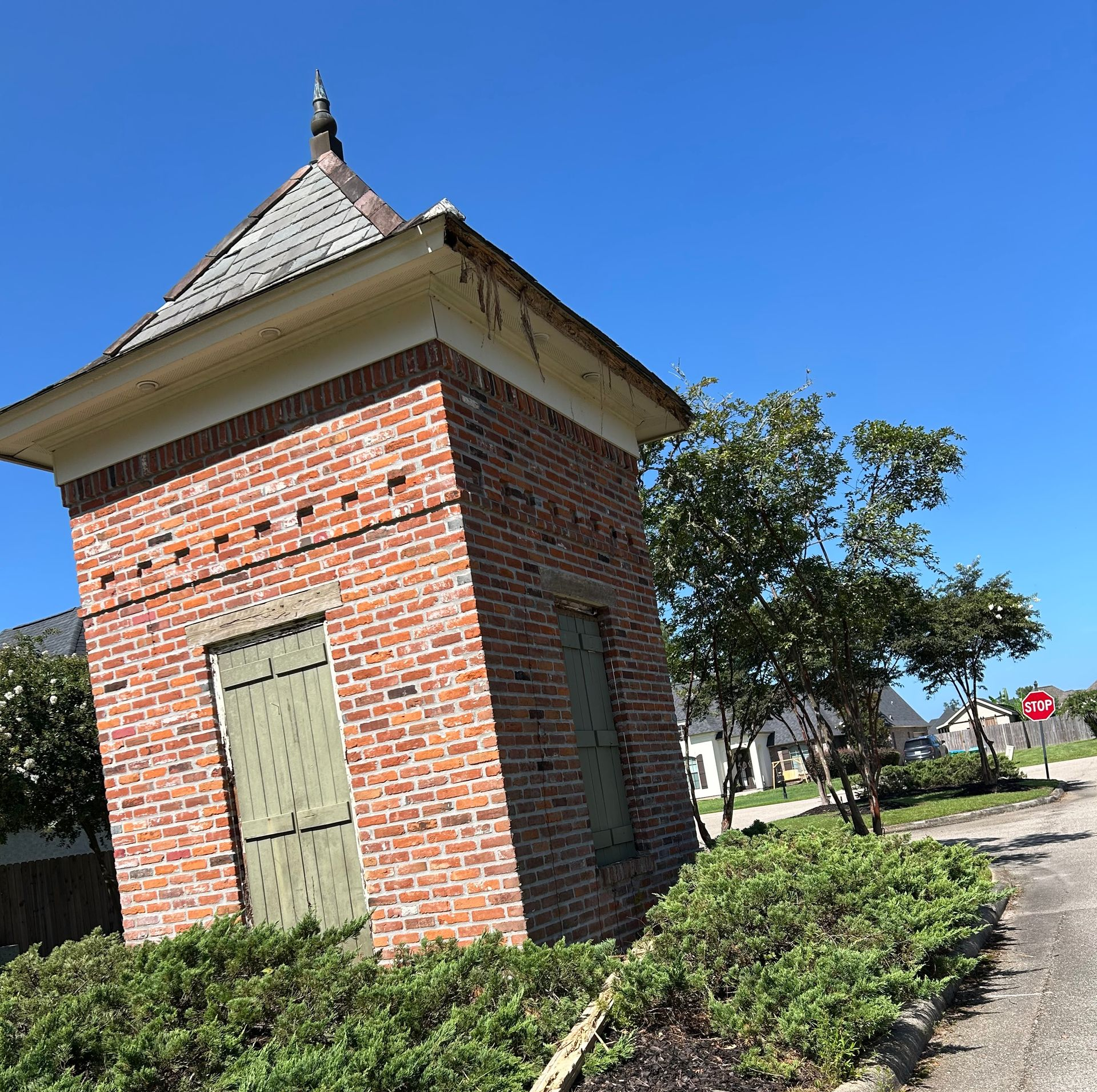 Small brick utility building with a metal roof beside a sidewalk and trees under a clear blue sky