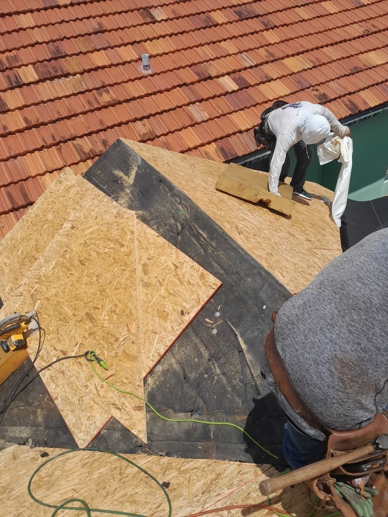 Workers repairing a shingled roof, with plywood patches and tools on a steep rooftop.