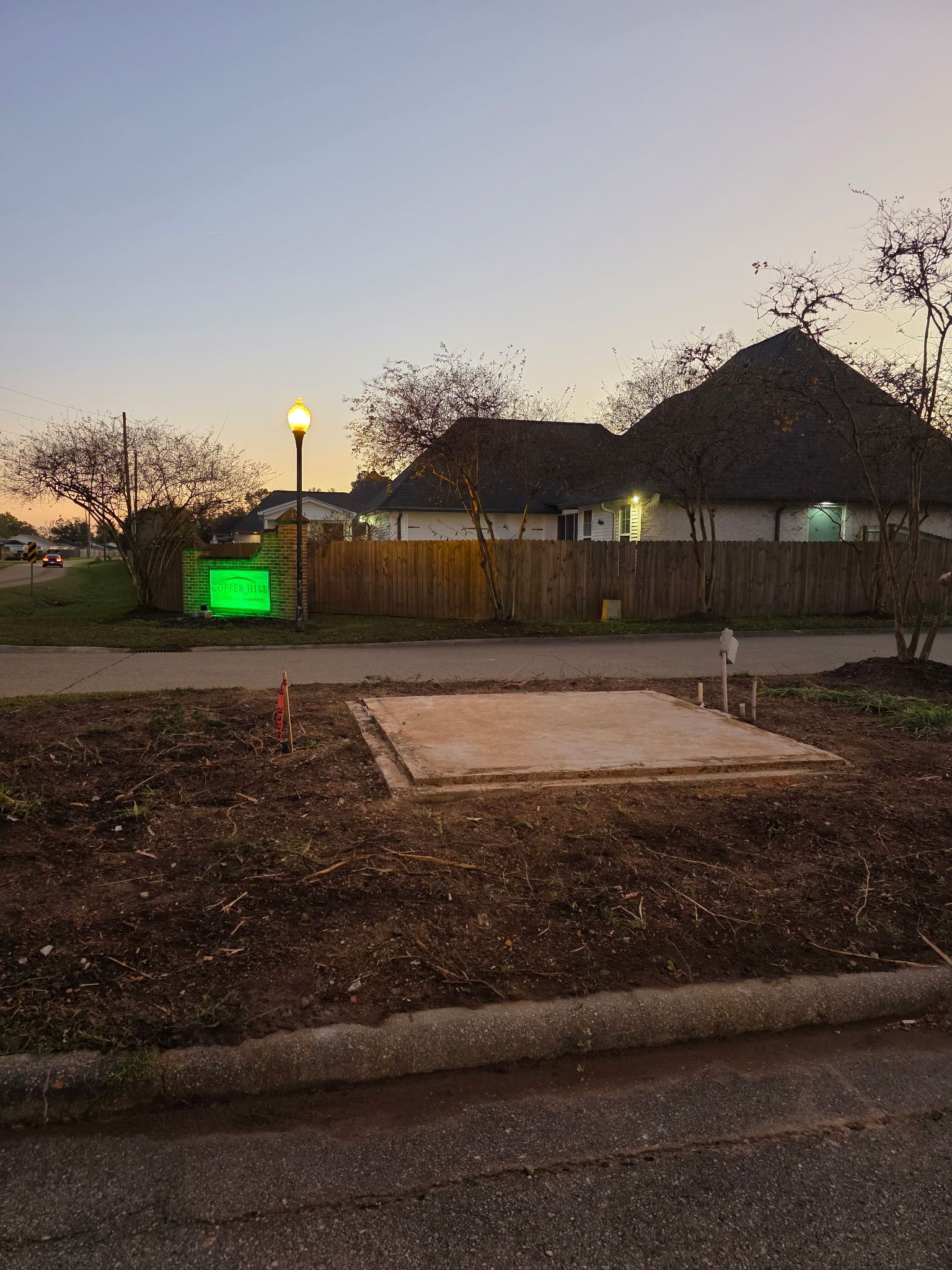 Residential lot at dusk with a vacant concrete pad, fenced yard, glowing green sign, and houses in the background