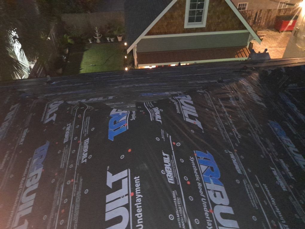 Night view of a rooftop with roofing underlayment, a dormer window, and lights from a nearby house.