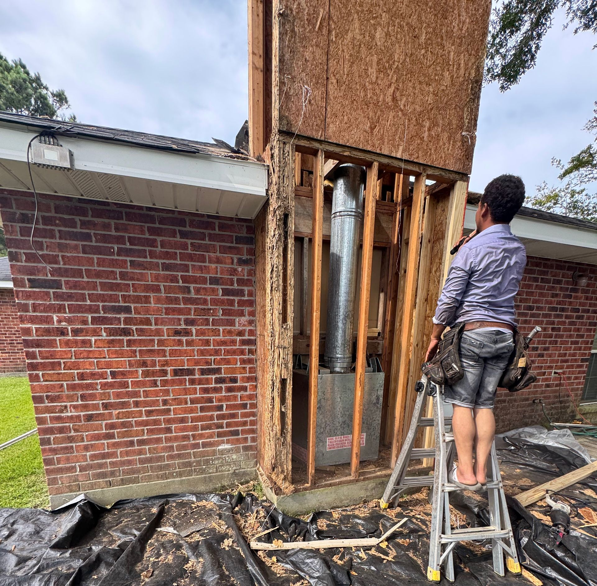 Worker on a ladder inspecting a damaged brick wall and exposed framing beside a tall rusted structure.