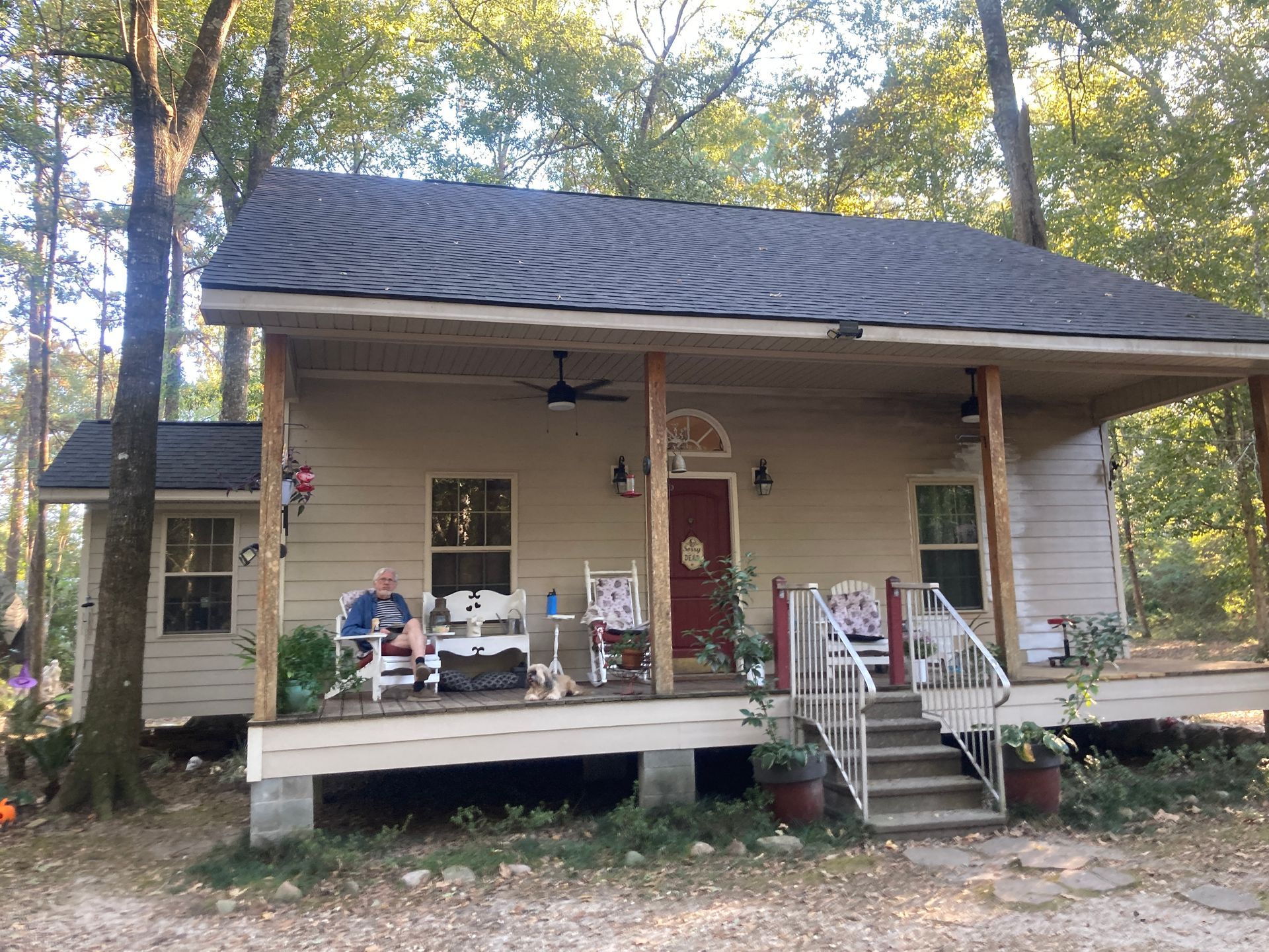 Small beige house with porch, steps, and two people sitting outside among trees.