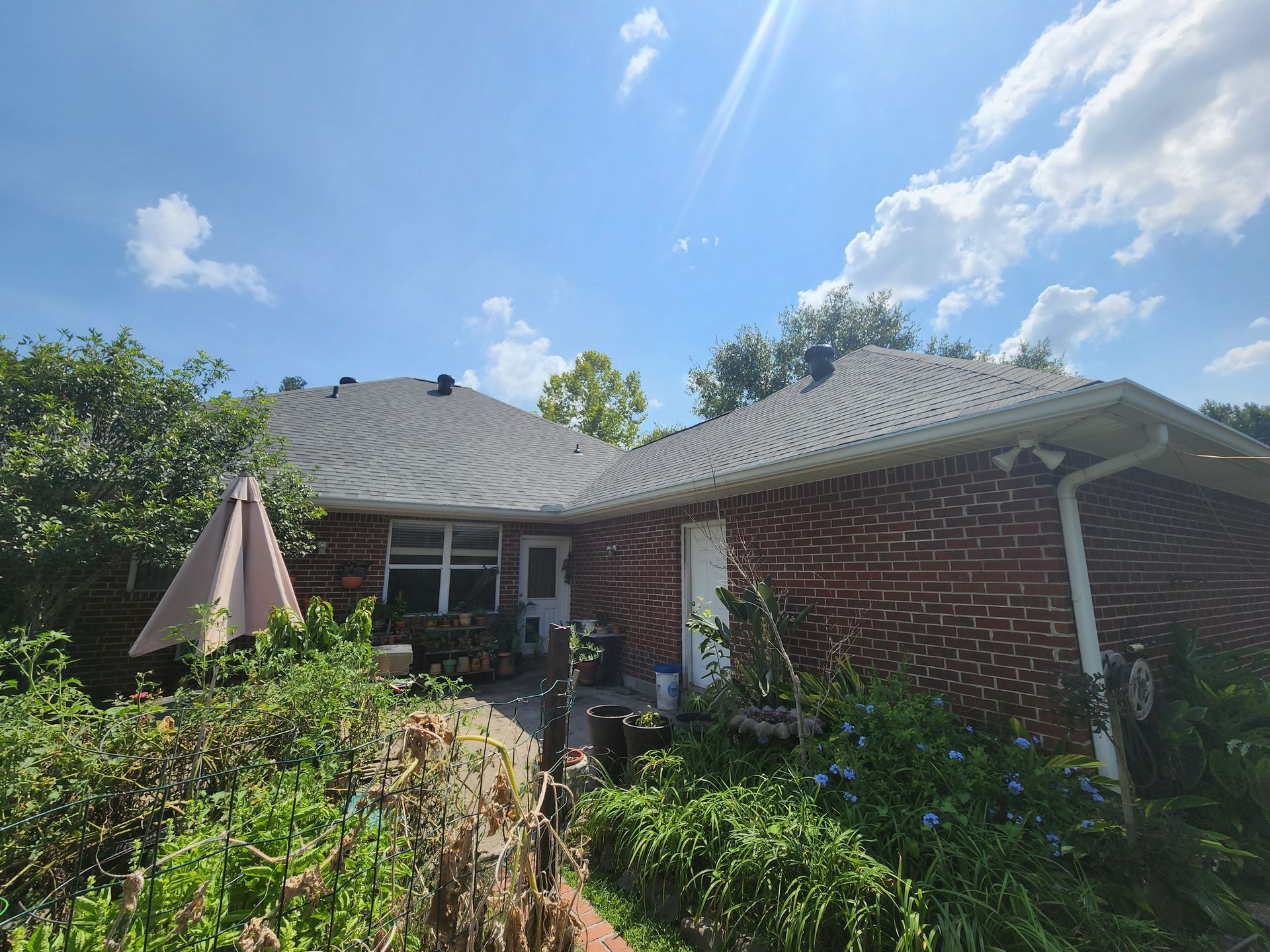 Single-story brick house with a shingled roof, front porch, and lush garden under a bright blue sky