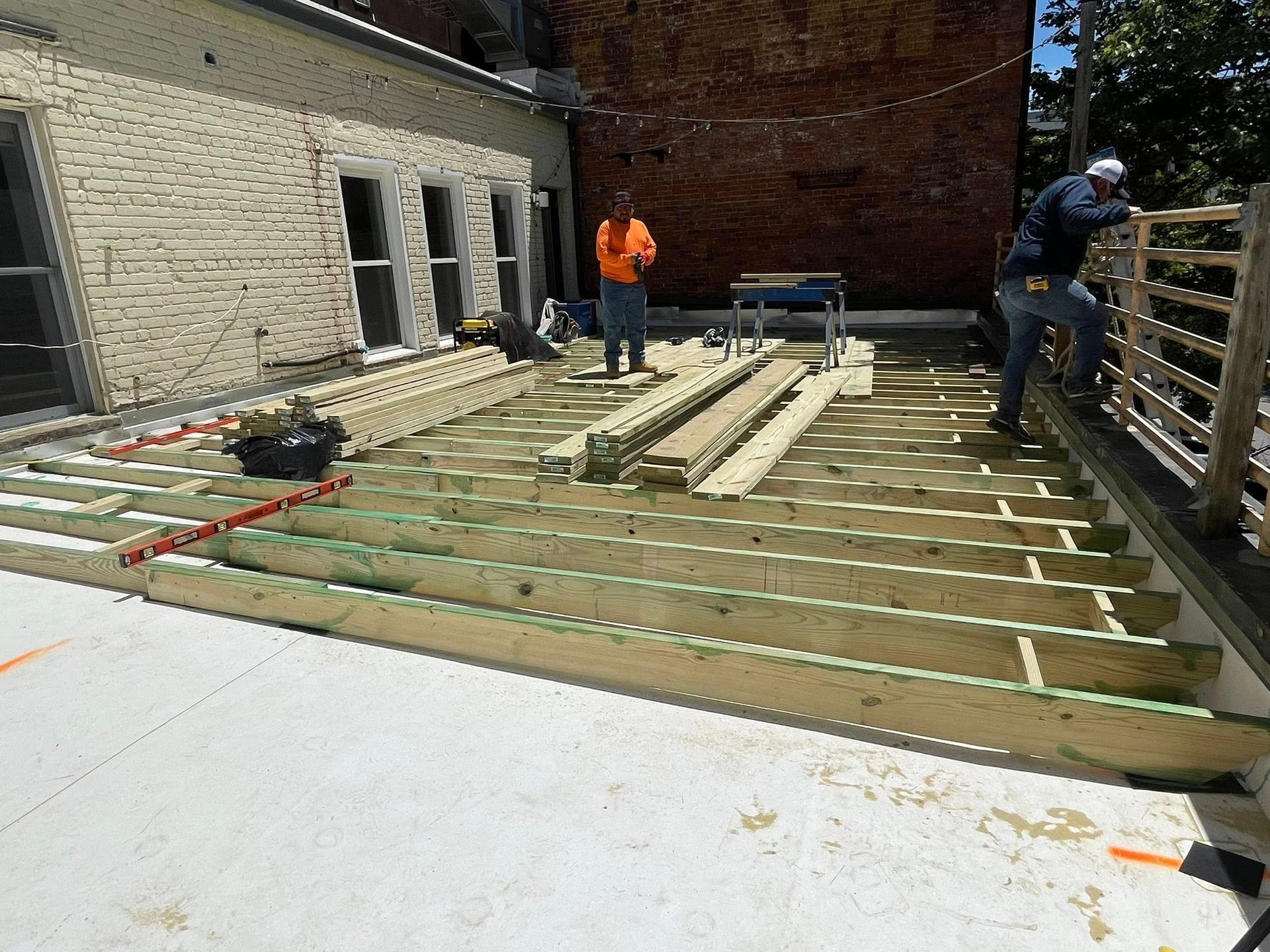 Workers installing wooden deck joists beside a brick house on a sunny rooftop terrace