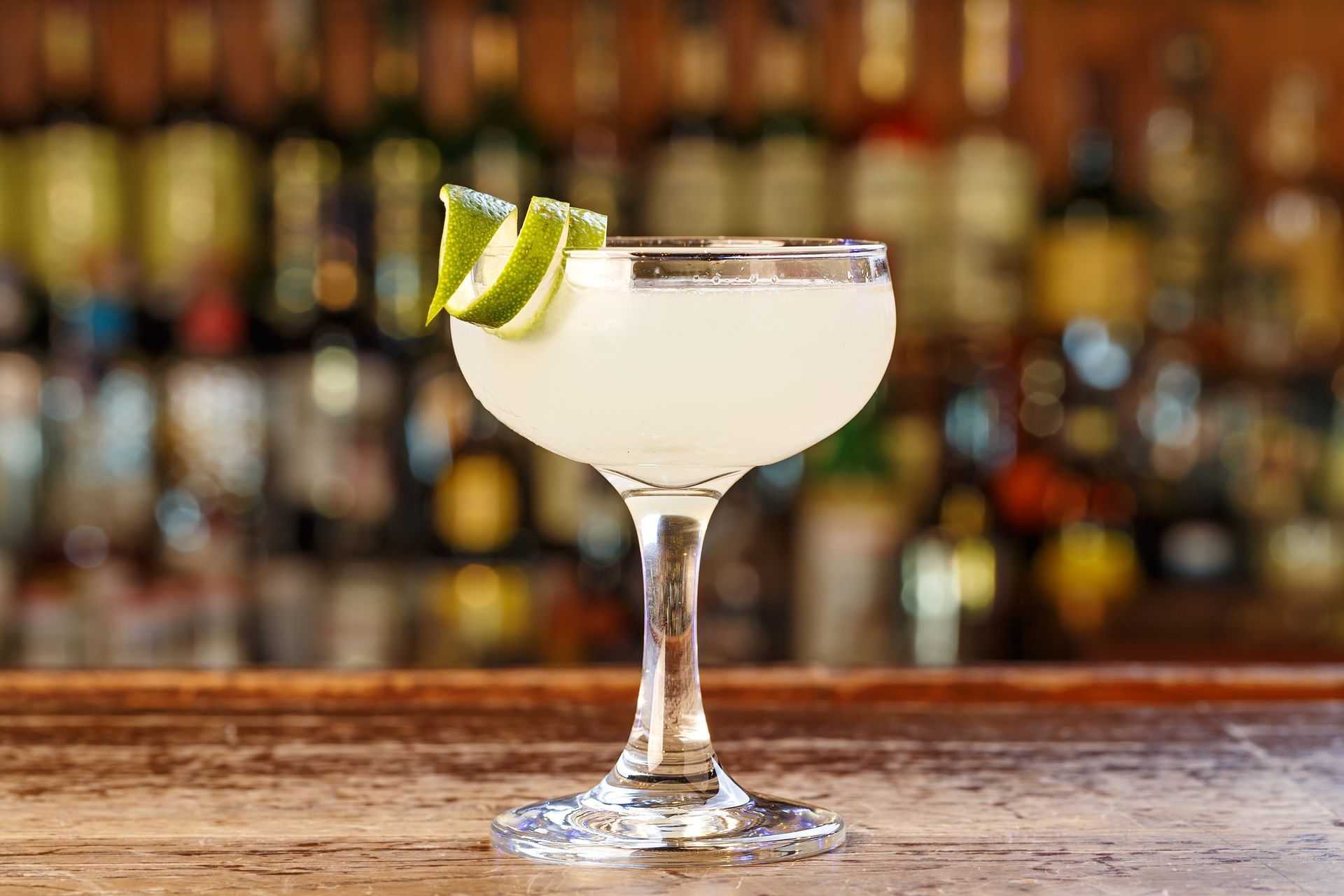 Cocktail in a coupe glass with a lime garnish on a wooden bar, bottles in the background.