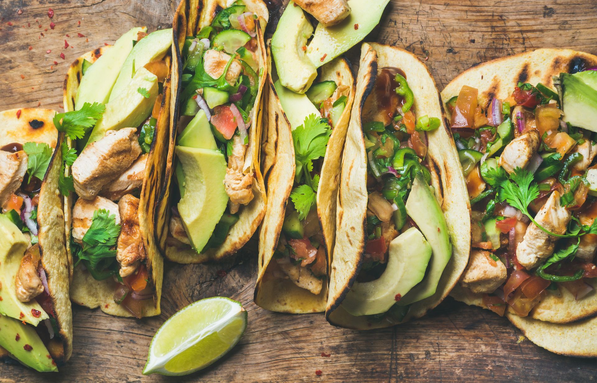 Grilled tacos with chicken, avocado, salsa, and lime on a wooden surface.