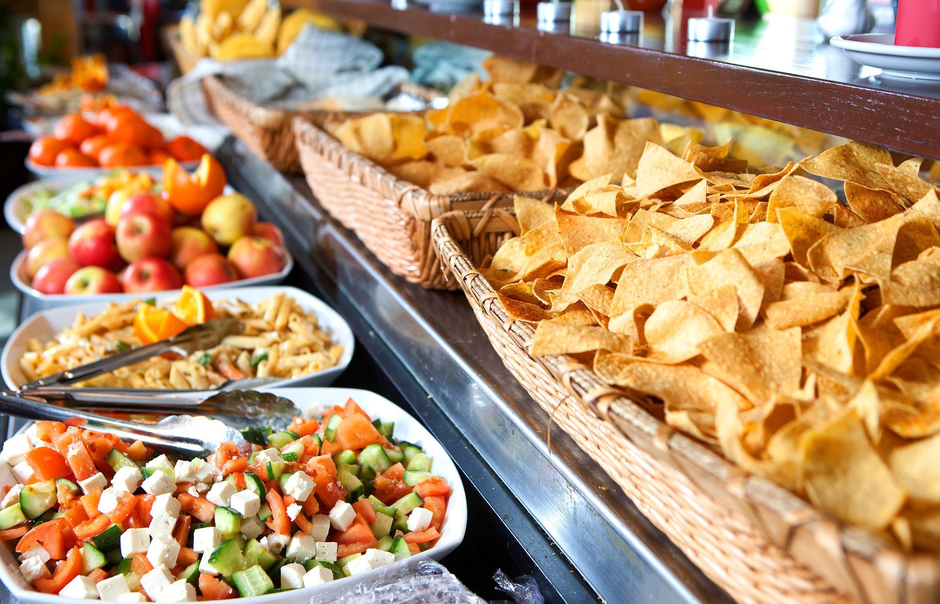 A buffet with various dishes: fruit, pasta salad, chopped vegetables, and tortilla chips.