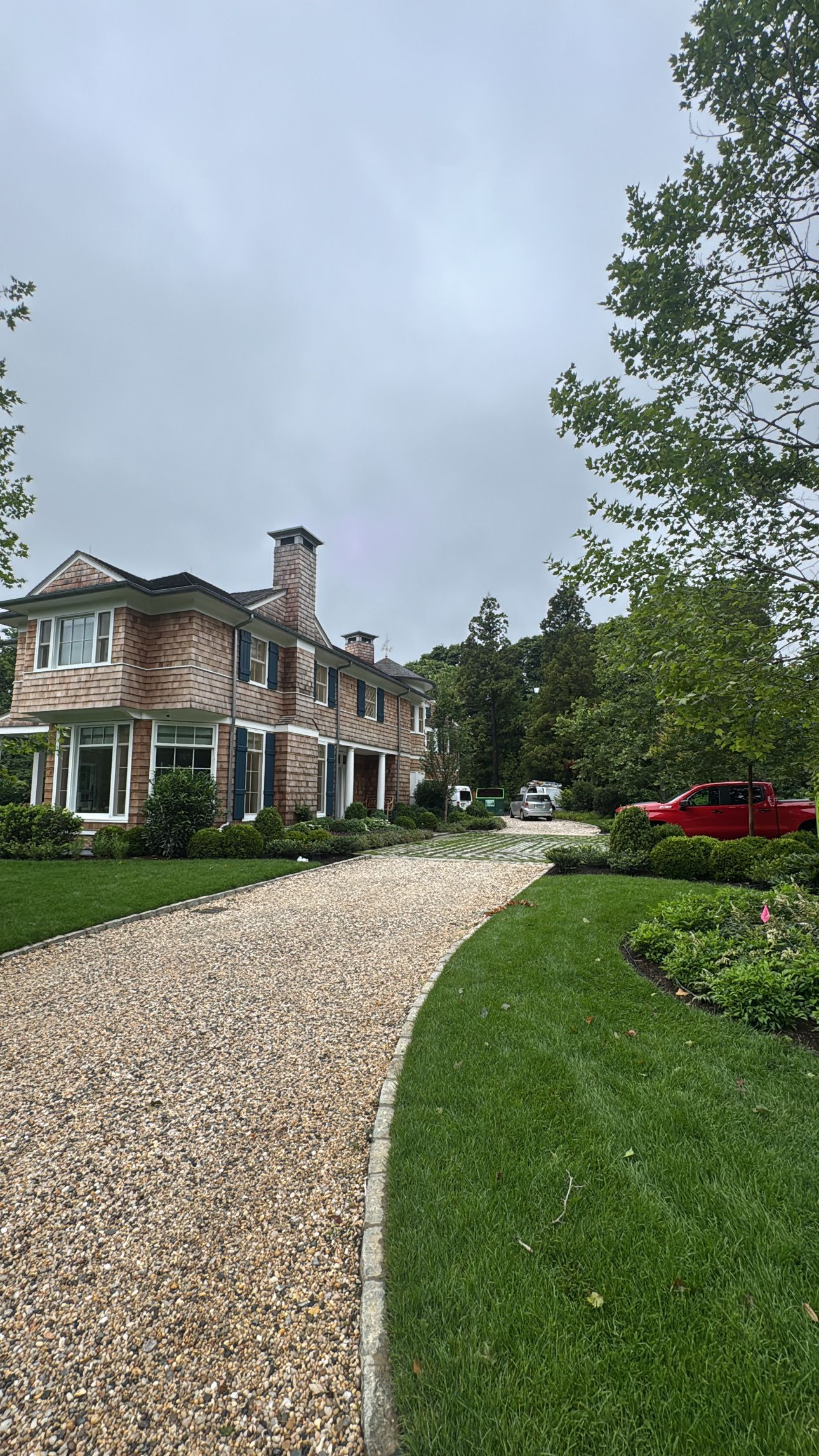 Curved gravel driveway leading to a large stone house with green lawn and trees under a cloudy sky
