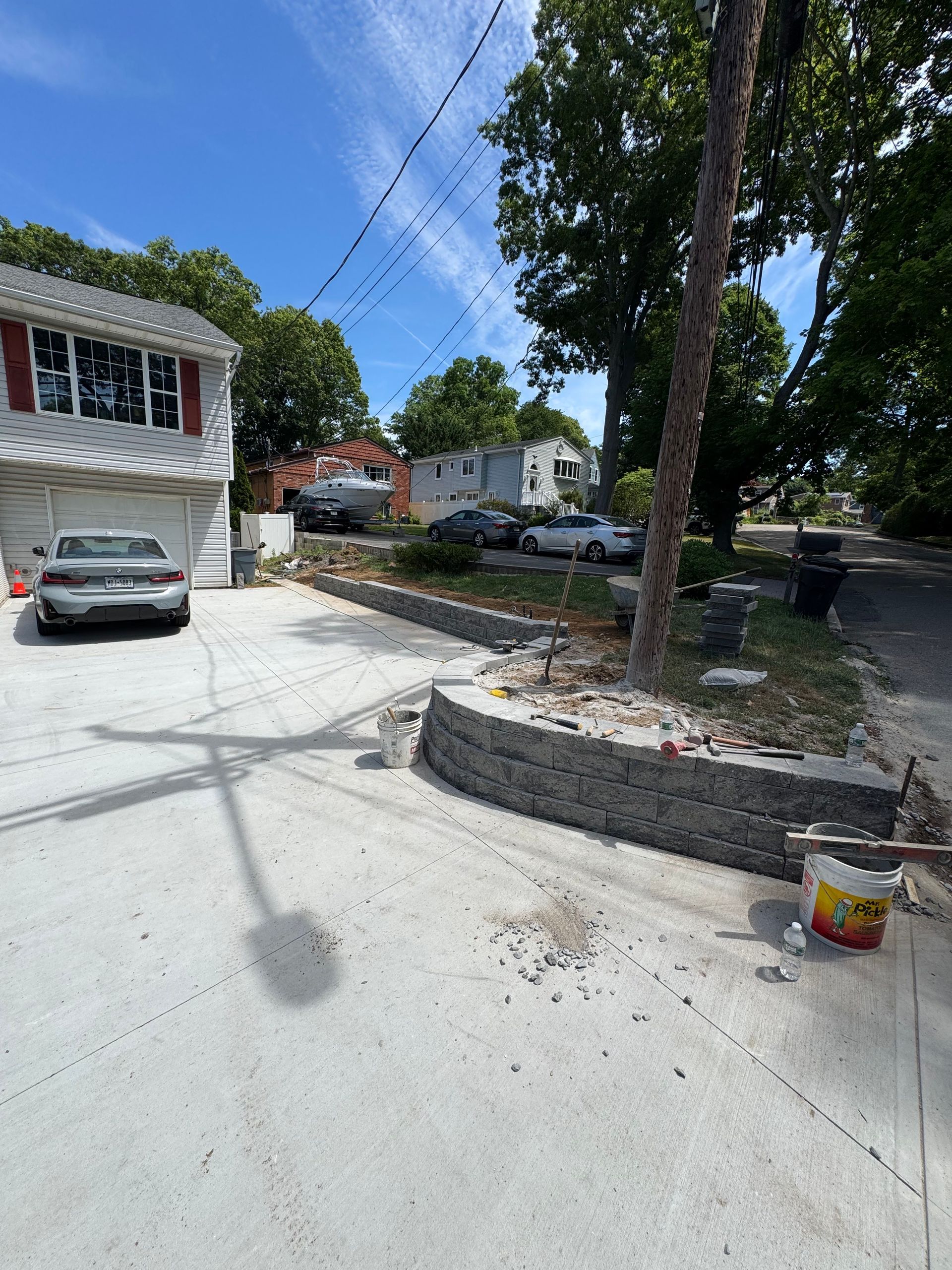 Driveway under construction with a parked car, stone retaining wall, trees, and a bucket of materials.