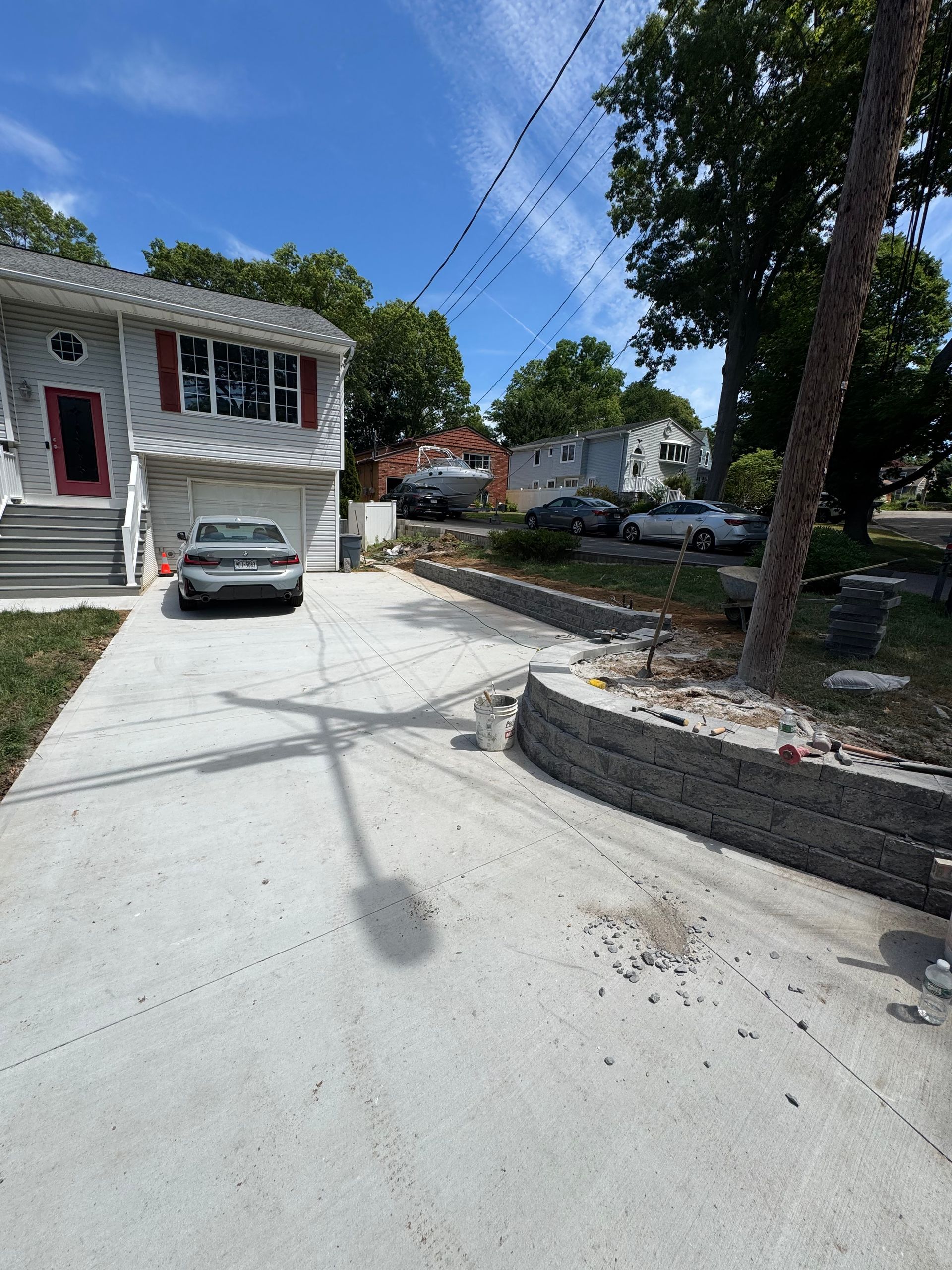 Driveway leading to a house with a parked car, stone retaining wall, and tree-lined residential street.