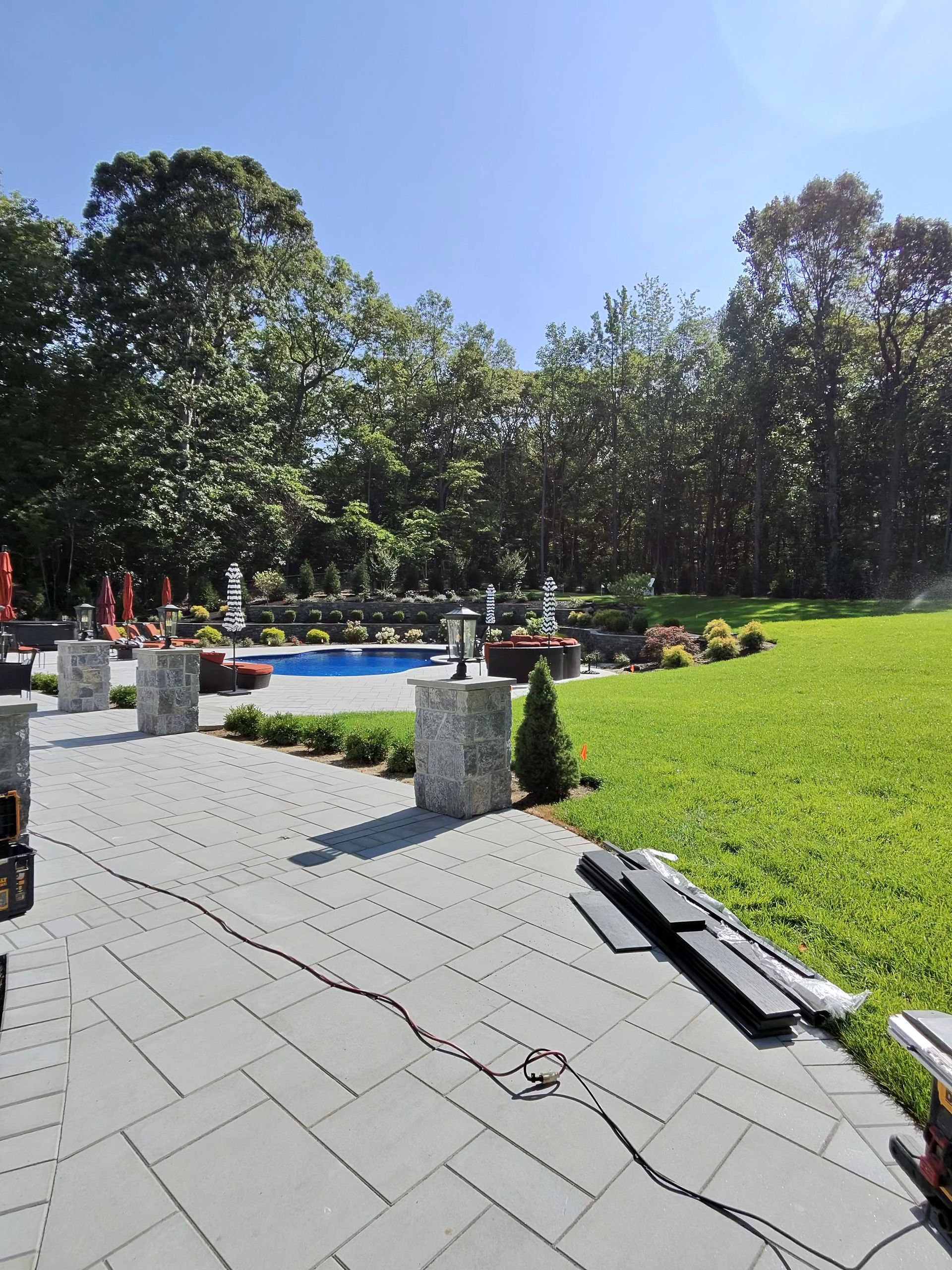 Sunny park walkway beside a bright green lawn, flowerbeds, and trees under a clear blue sky.
