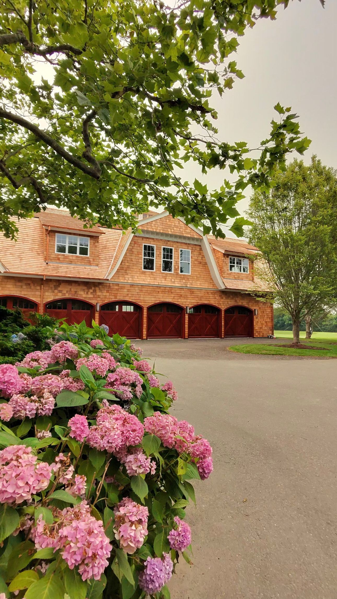 Pink hydrangeas in front of a red-roofed house with arched wooden garage doors under leafy trees