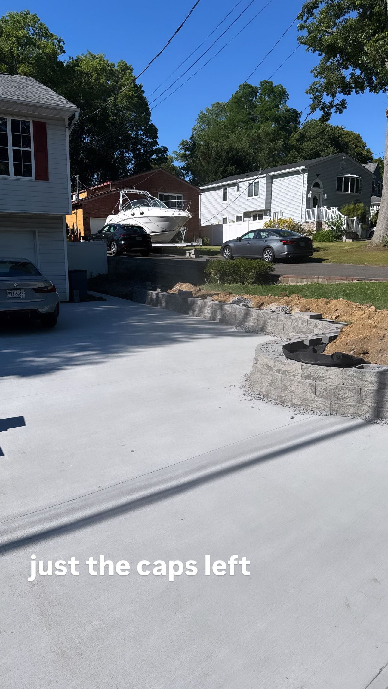 Concrete driveway in a suburban neighborhood with houses, trees, parked cars, and stone landscaping.