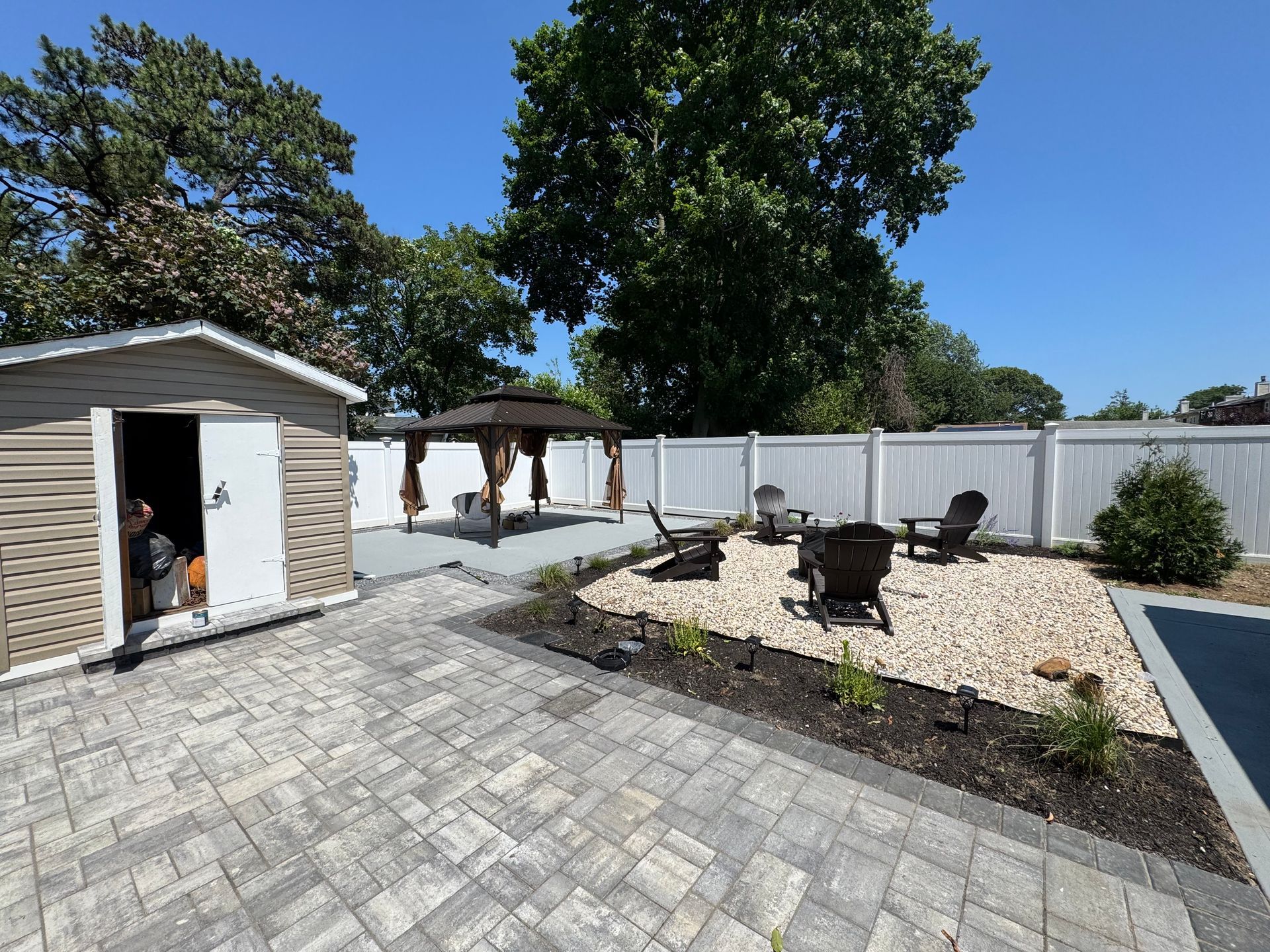 Backyard patio with pavers, fire pit, pergola, white fence, and a shed under blue sky