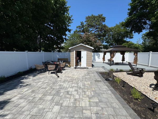 Backyard patio with paver walkway, shed, gazebo, grill, and white fence under a sunny blue sky