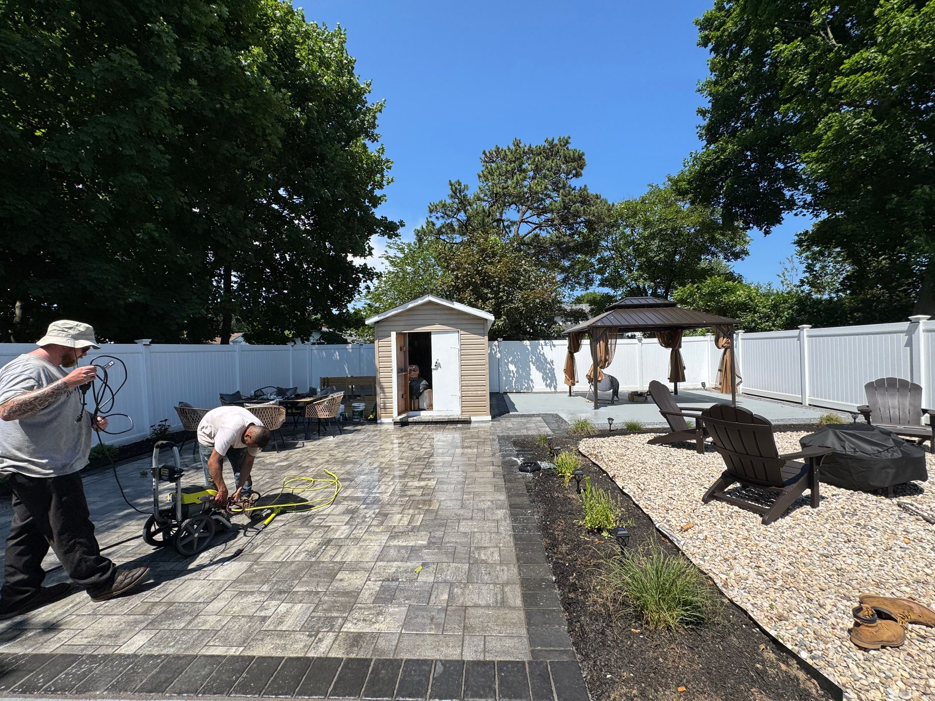 People working in a paved backyard garden with raised beds, trees, and sheds on a sunny day