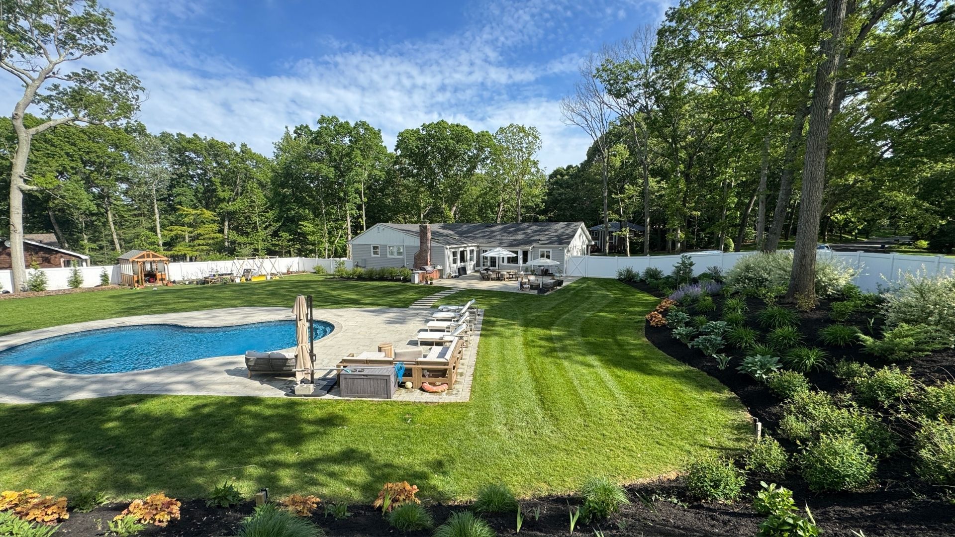 Backyard pool with lounge chairs, green lawn, flower bed, and a patio beside a white fence
