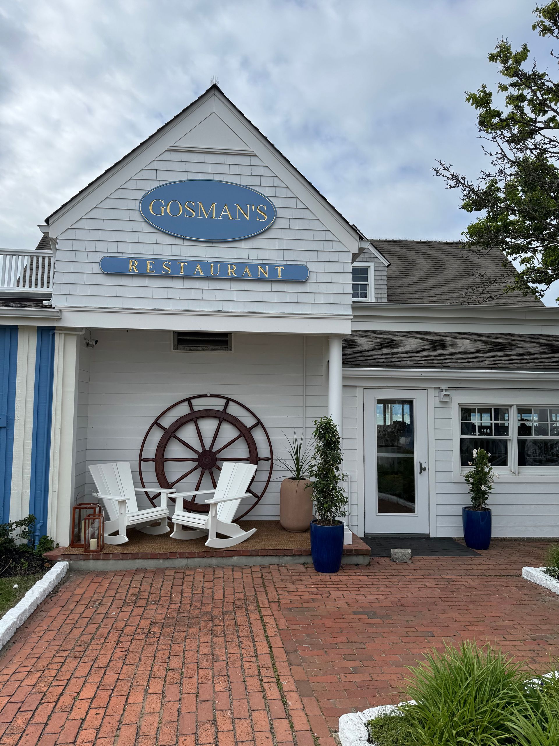 White seaside storefront with blue sign, brick walkway, wheel decor, and potted plants