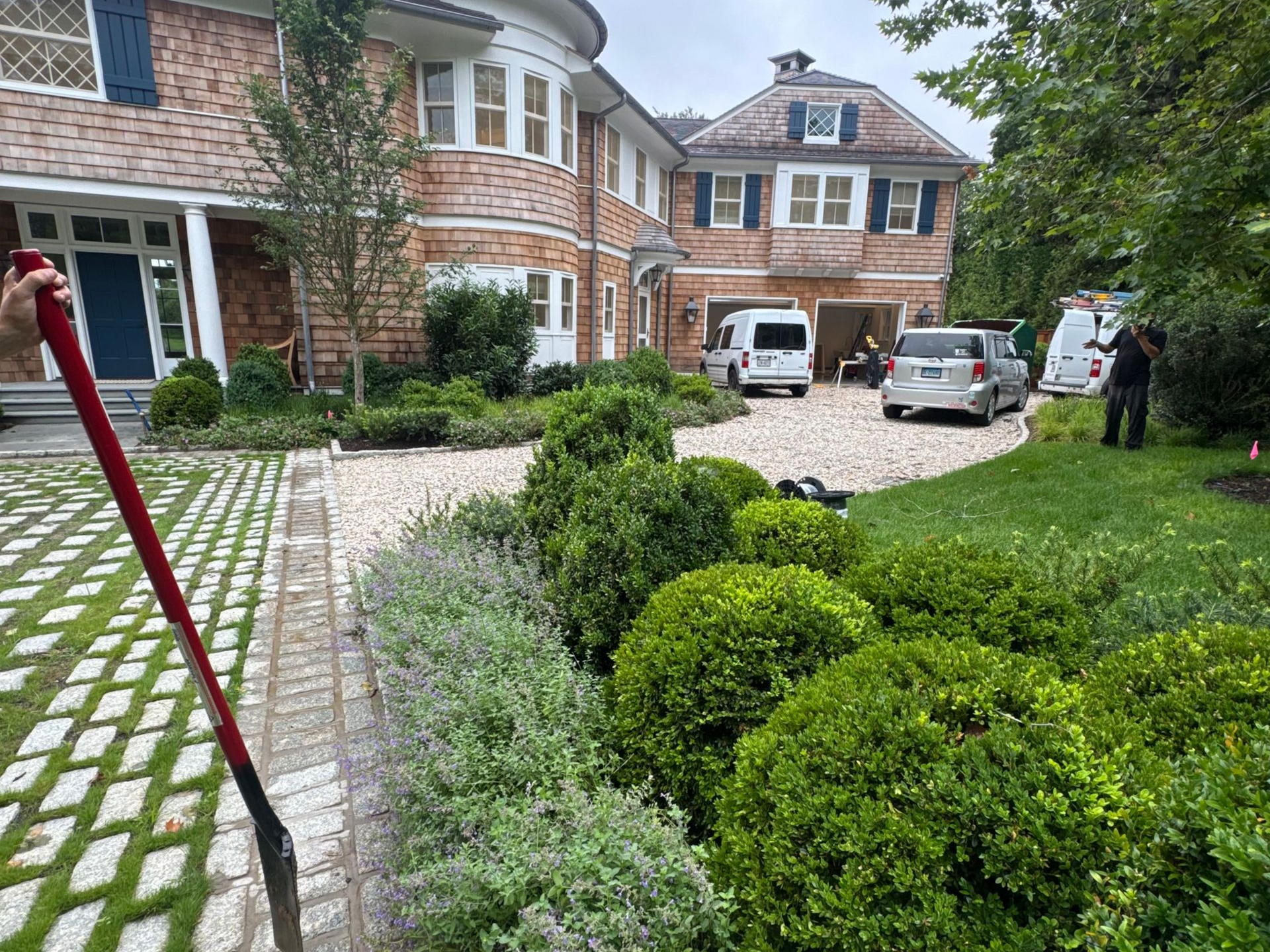 Brick house driveway with parked cars, manicured shrubs, and a paved garden path leading to the entrance