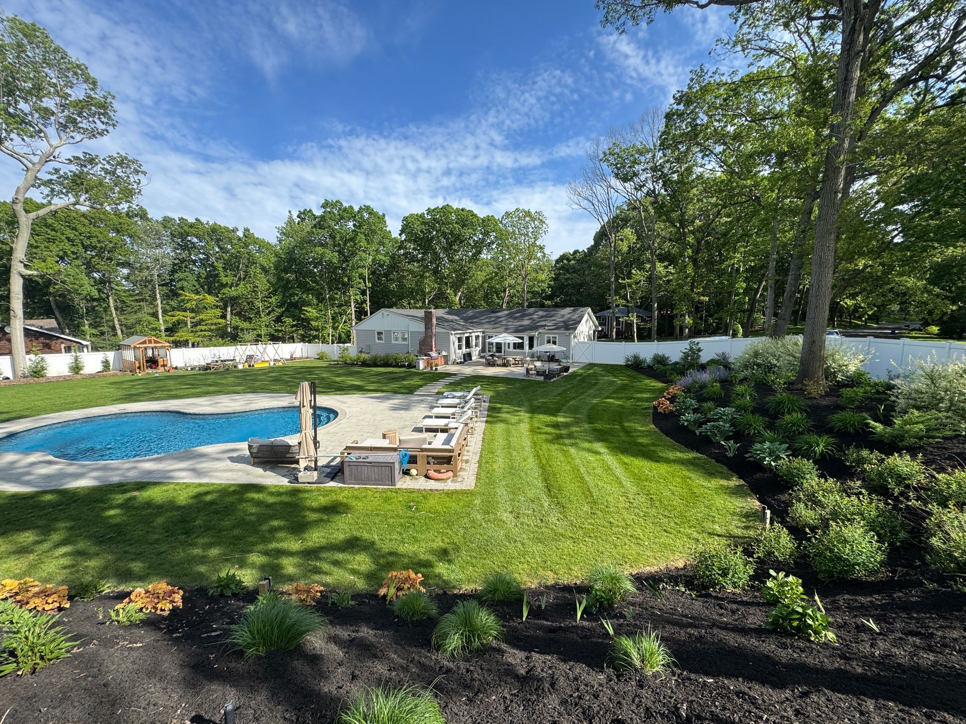 Backyard with a swimming pool, lounge chairs, green lawn, and trees under a blue sky