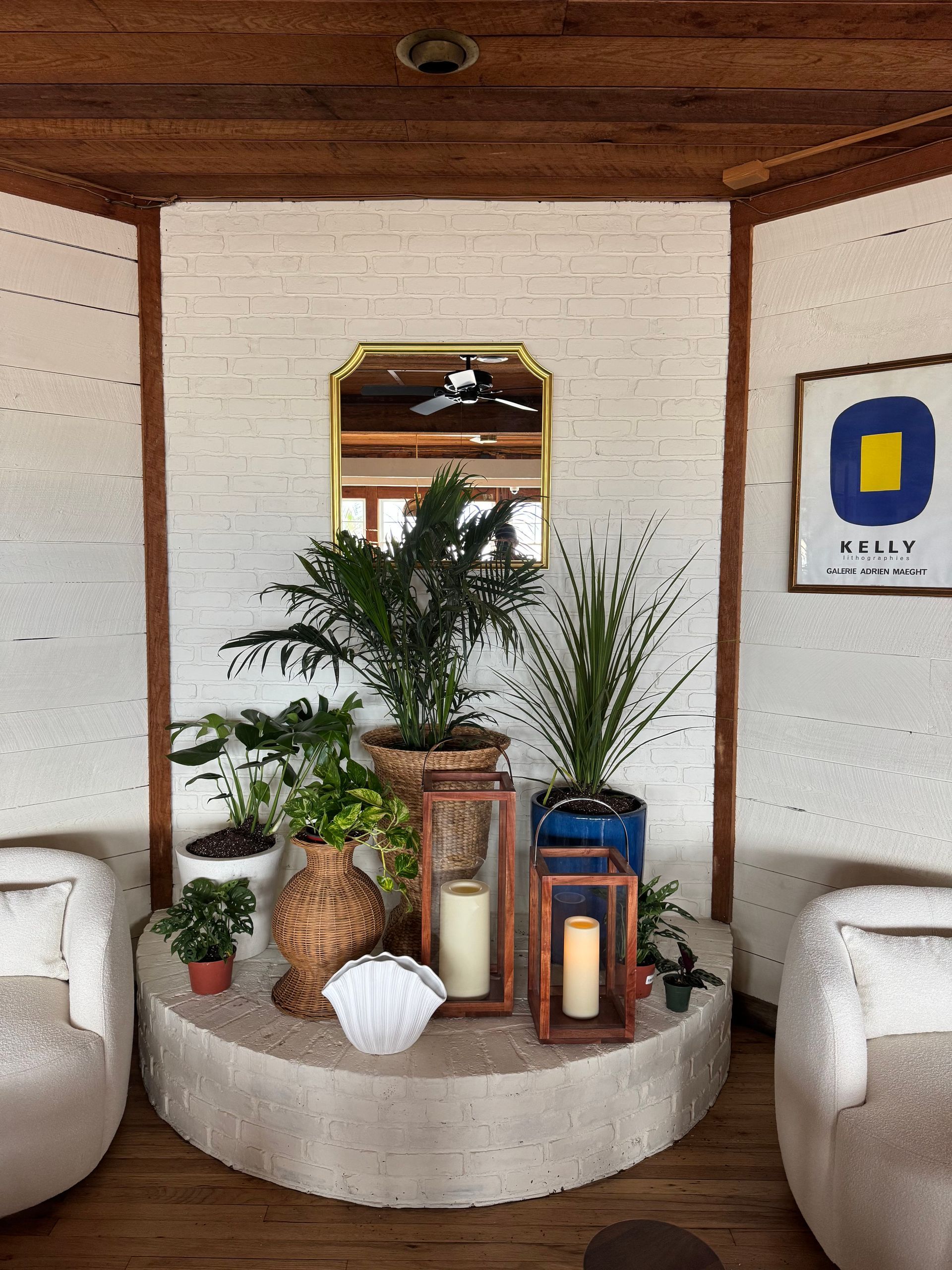 Potted plants and lanterns on a round white pedestal in a cozy white-and-wood room with a mirror and wall art.