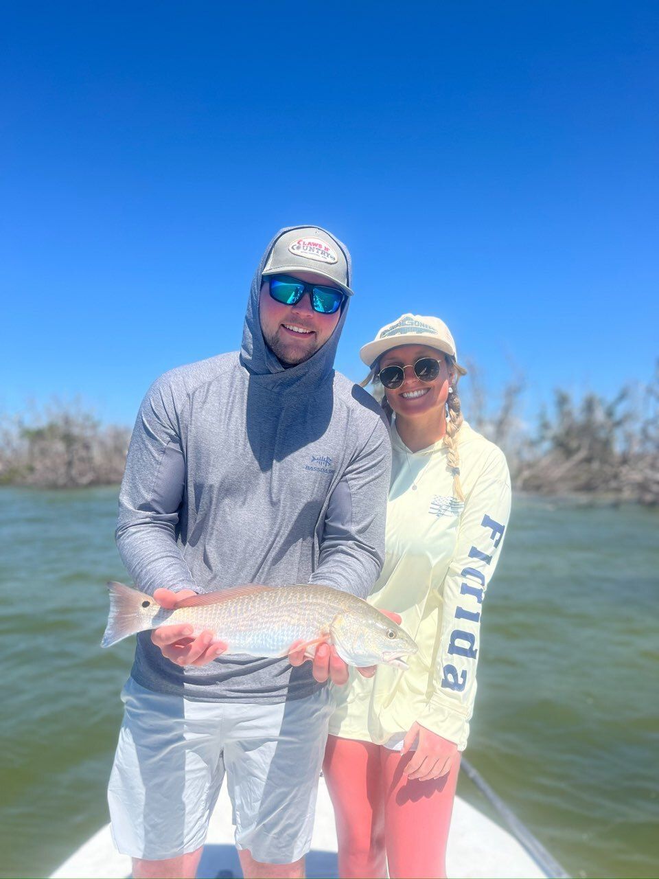 A man and woman on a boat hold up a redfish they caught. They are smiling, with blue skies and water.