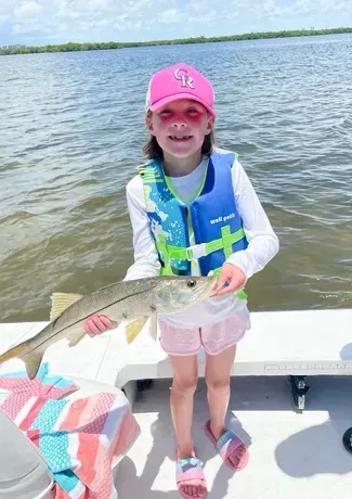 A young girl in pink hat and life vest holds a snook fish on a boat. Overcast day, near the water.