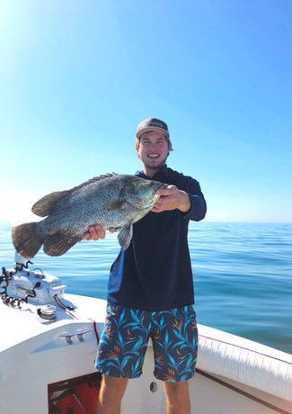 Man on a boat smiles, holding a large gray fish against a bright blue sky and water. He wears a blue shirt and patterned shorts.