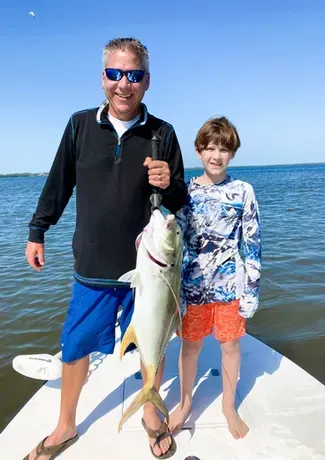 Man and child on a boat, holding up a large yellow fish. Man smiles, wearing blue shorts and sunglasses; child smiles, wearing orange shorts.