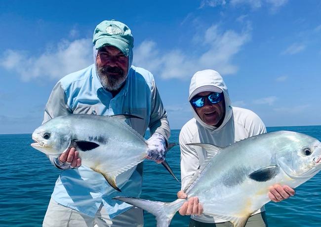 Two men on a boat holding up large, silver fish they've caught. They're smiling, with blue sky and water in the background.